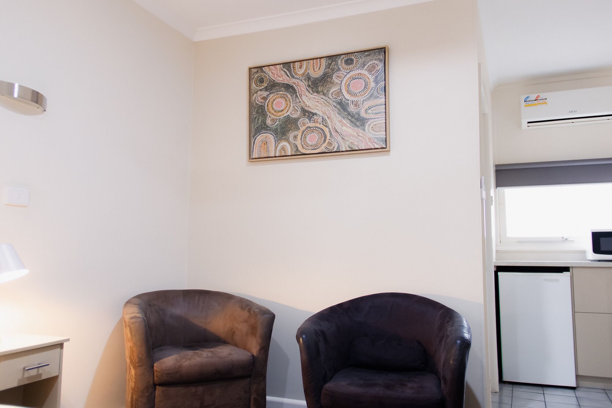 A room with two leather chairs, a wall-mounted artwork, a white desk, and a kitchenette with an air conditioner.