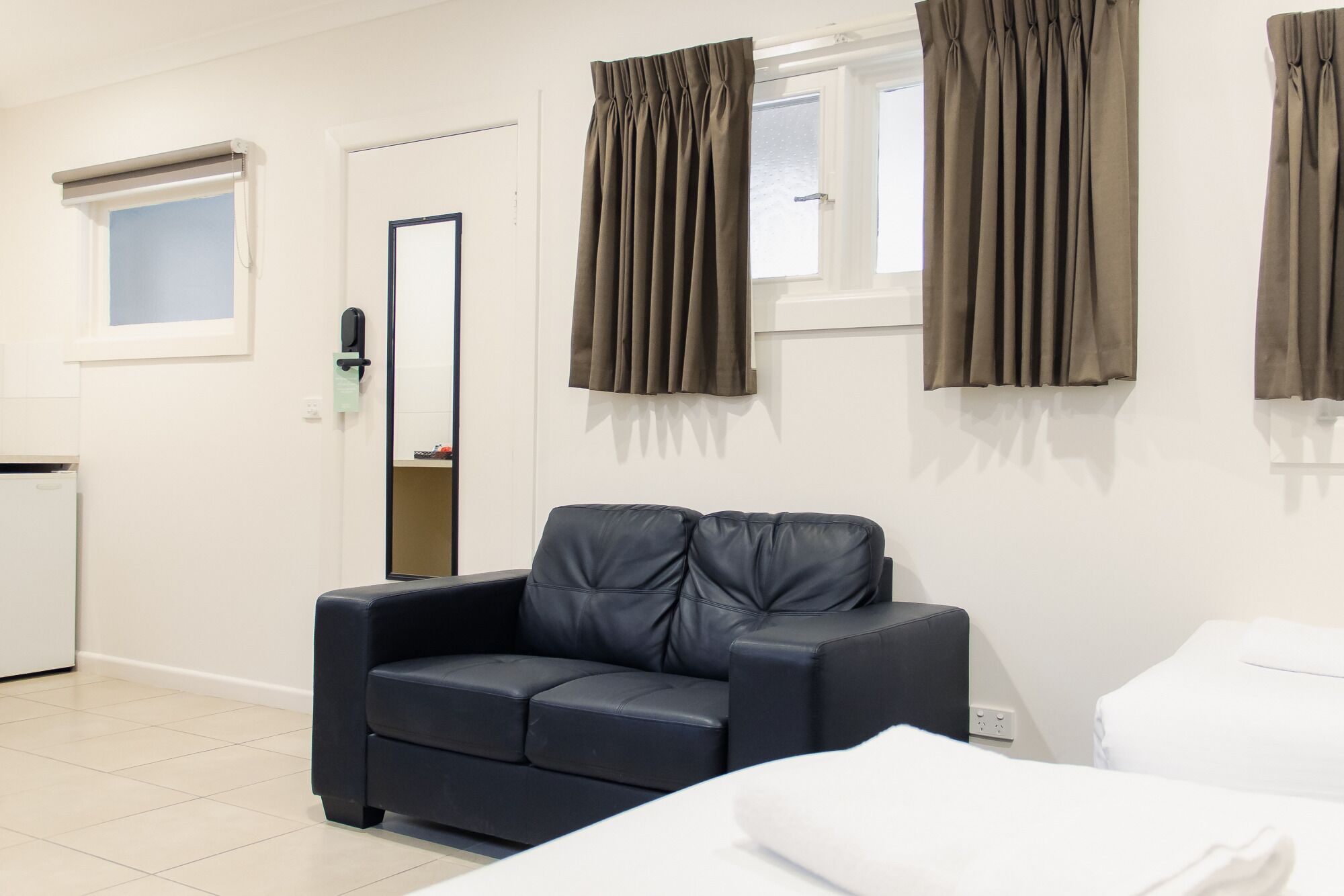 A hotel room with a dark leather sofa, a bed with white linens, and two windows with brown curtains.
