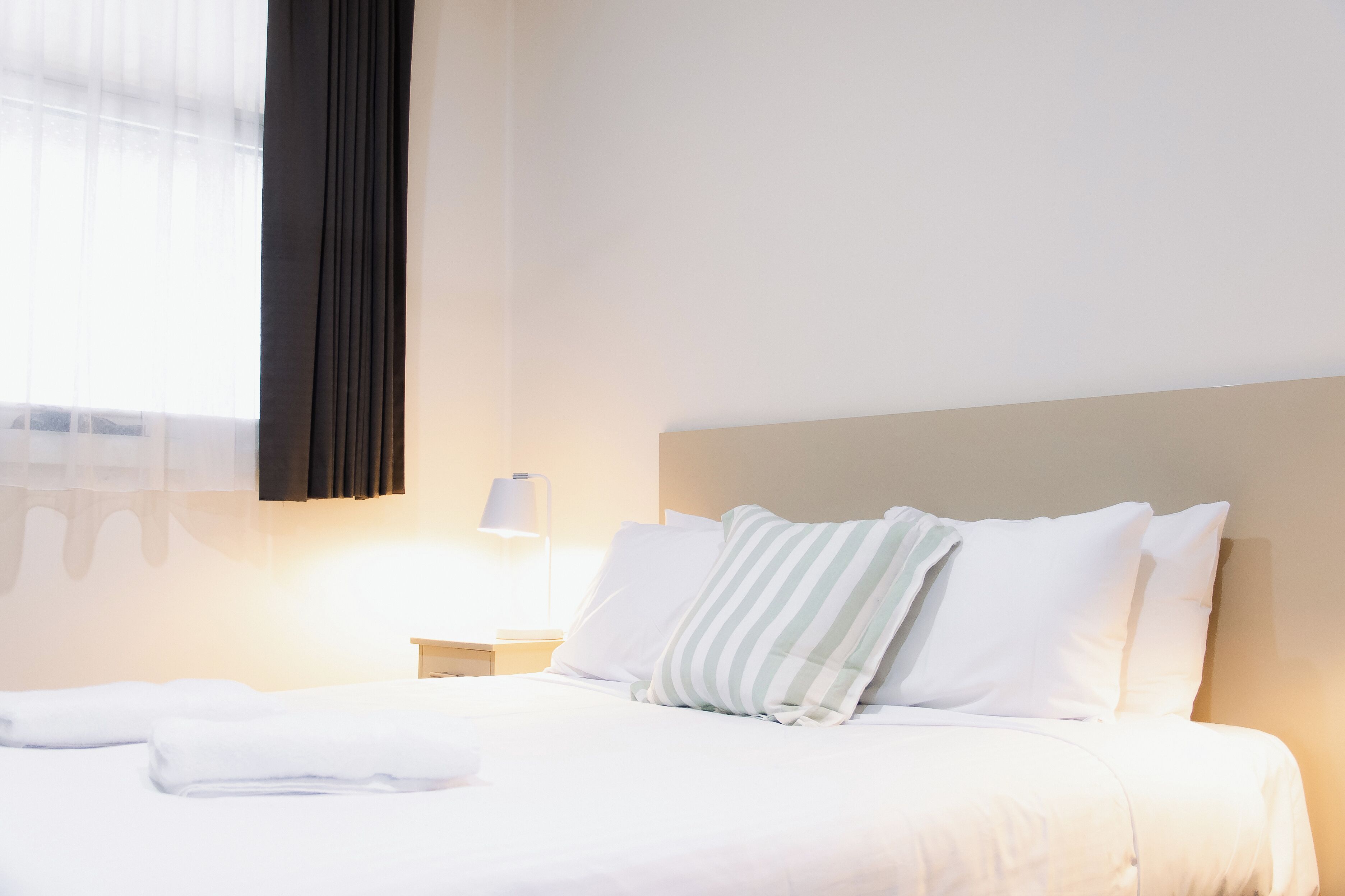 Un lit bien fait, avec du linge de lit blanc et des oreillers à rayures, une lampe de chevet et une fenêtre ornée de rideaux légers.