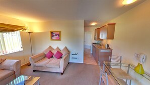 A cozy living room with a sofa, a small table, and a kitchen area with cabinets and a sink. - Tresori Motor Lodge (Akaroa)