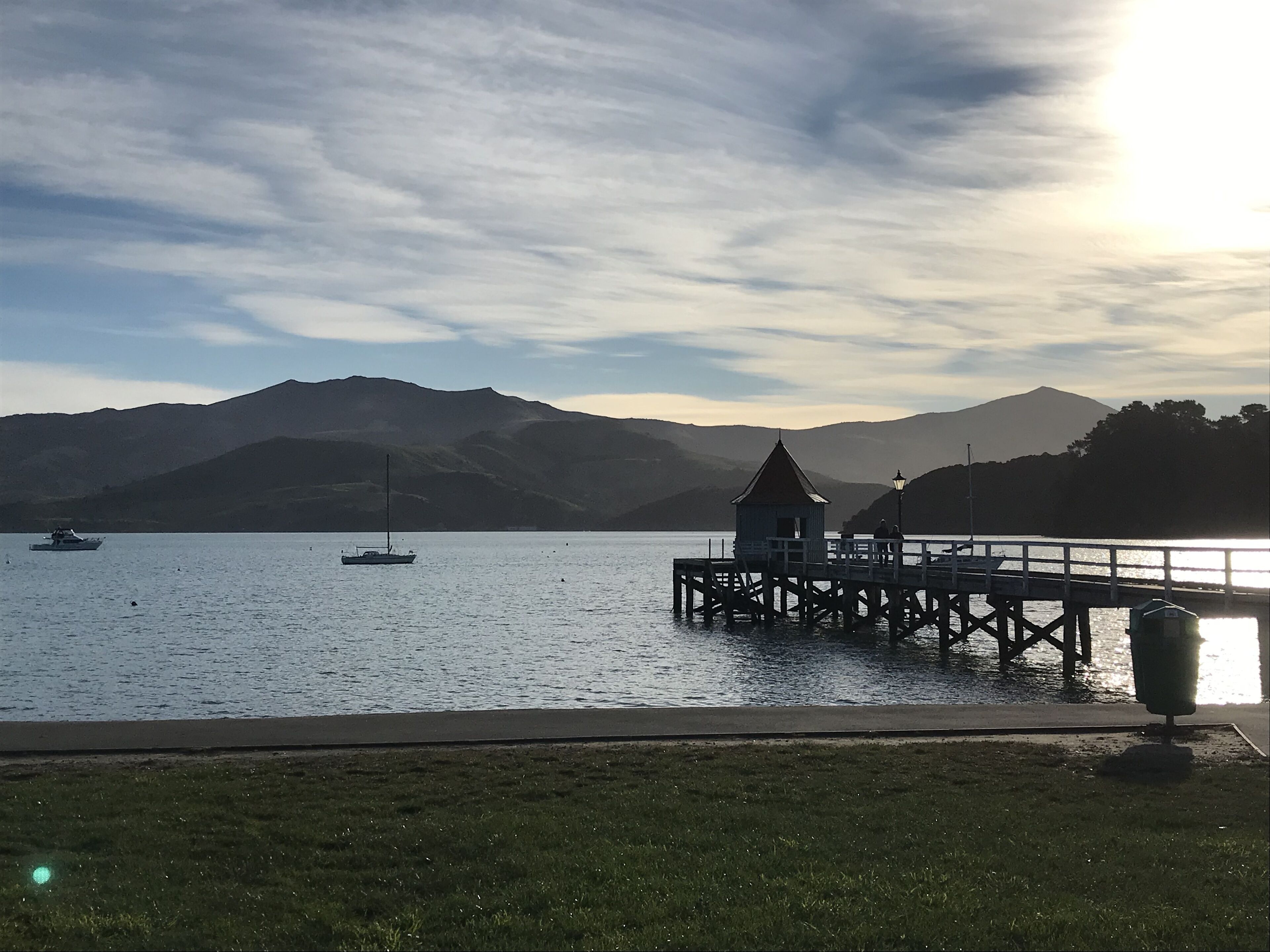Une jetée s’avançant dans une étendue d’eau calme, avec des bateaux, une petite construction et des montagnes en arrière-plan.