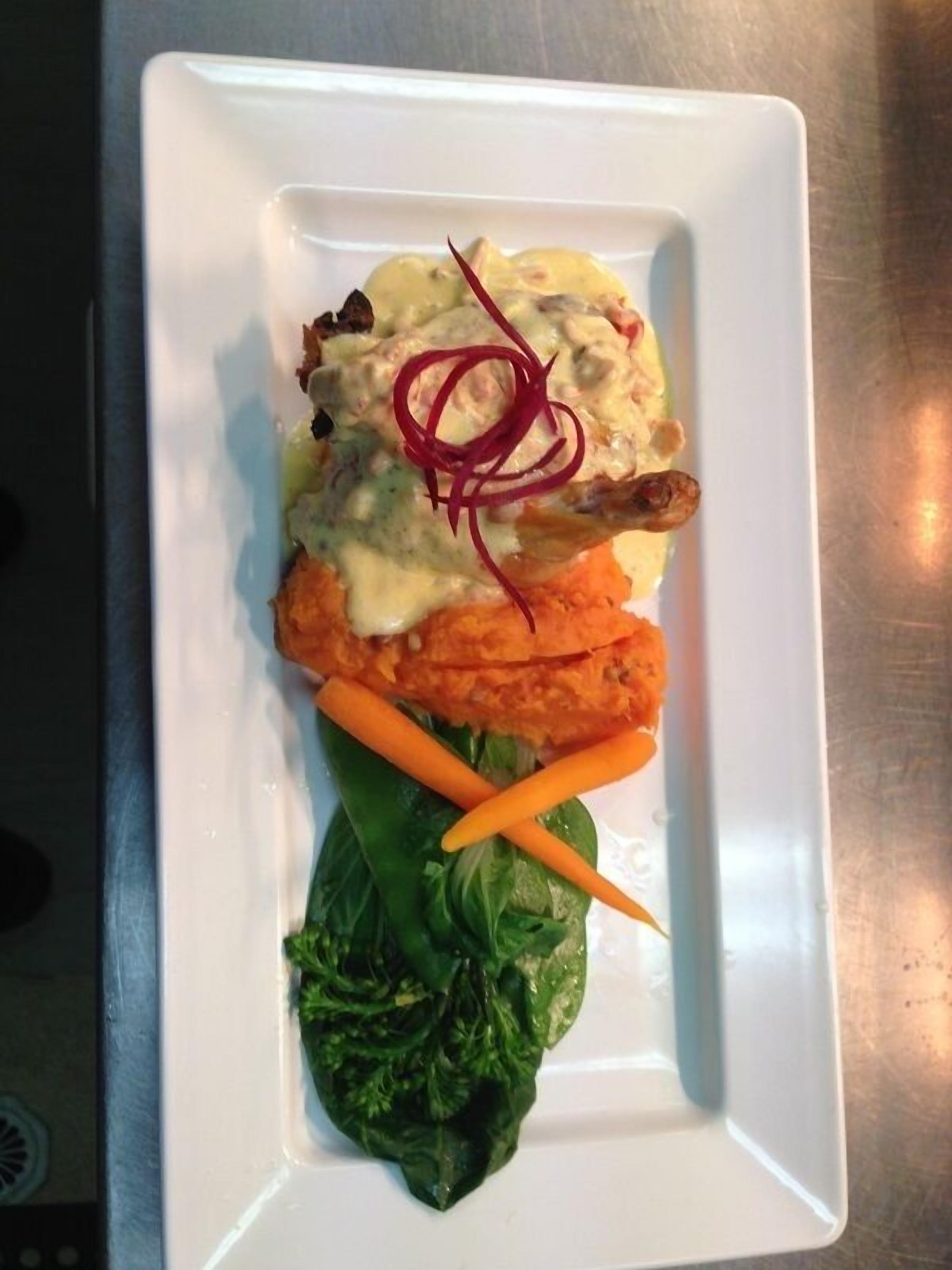 A white rectangular plate with a piece of meat topped with a creamy sauce, two orange carrot sticks, and a bed of green leafy vegetables.