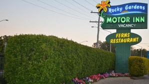 Un letrero de motel con los nombres 'Rocky Gardens Motor Inn' y 'Fern's Restaurant', rodeado de un seto bien cuidado y flores coloridas.