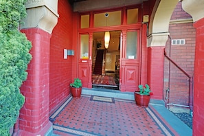 A red brick building entrance with a welcome mat, two potted plants, and a patterned floor. - Edinburgh Gallery Bed & Breakfast (Hobart)