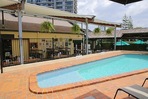 A swimming pool with a tiled edge, surrounded by a brick patio. There are outdoor tables and chairs, and a covered area with a roof. - Darcy Arms Motel (Surfers Paradise)