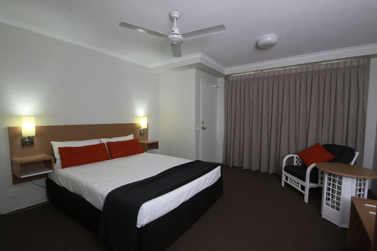 Una camera d'albergo con un letto, una sedia, un tavolo, un ventilatore a soffitto e una porta.