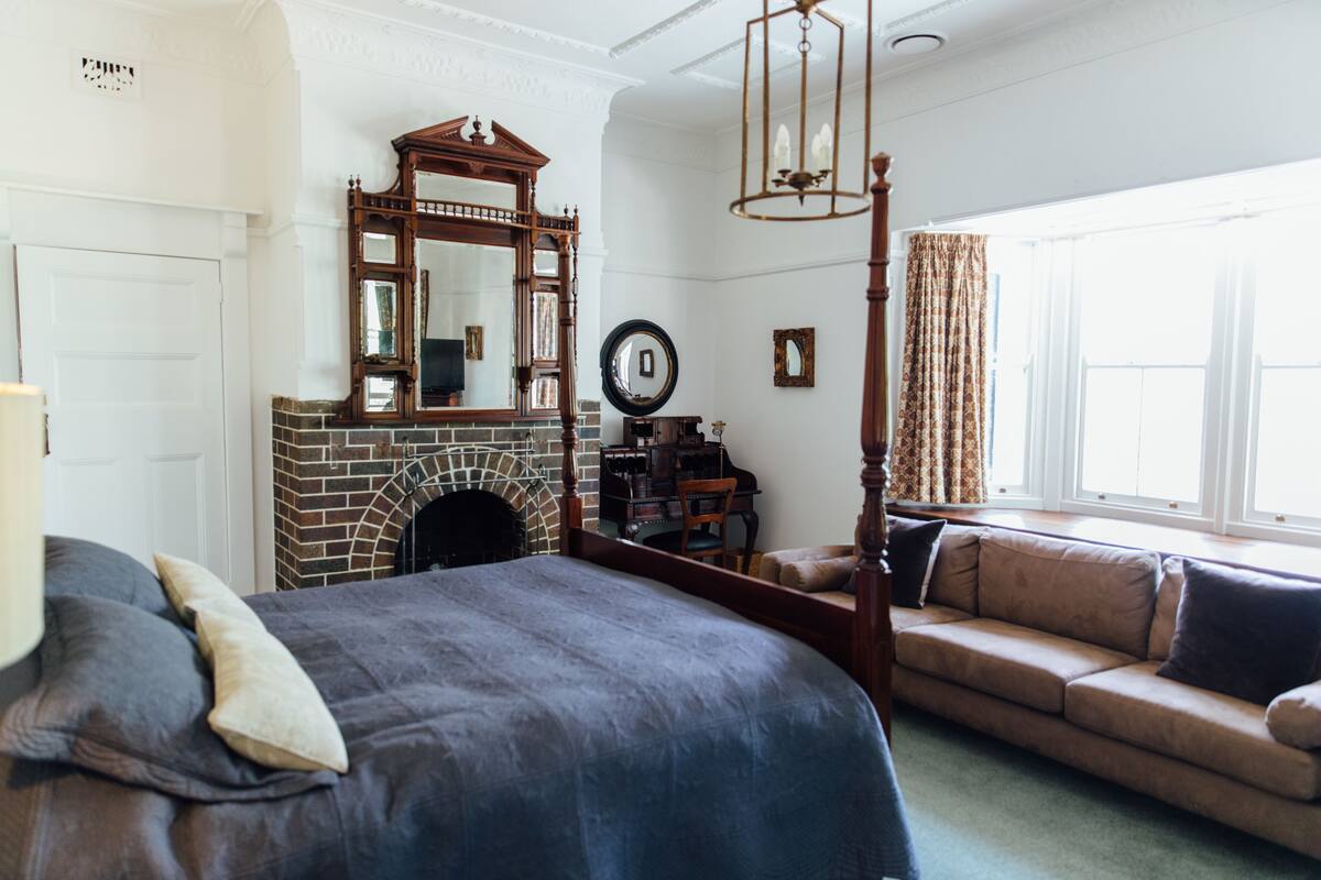 A bedroom with a four-poster bed, a fireplace, a large mirror, a sofa, and a window with curtains.