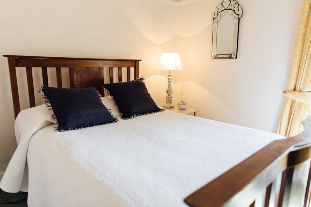 A neatly made bed with a white duvet and two dark pillows, a wooden headboard, a bedside lamp, and a mirror on the wall.
