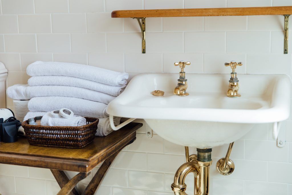 A bathroom with a white sink, two gold faucets, a wooden shelf above the sink, and a wicker basket with towels on a wooden table.