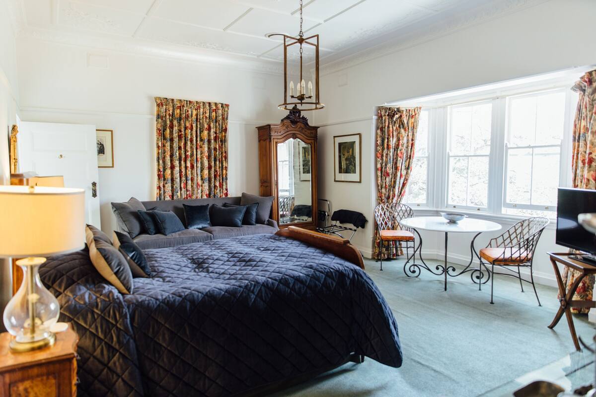 A bedroom with a large bed, a desk, and a chair. There is a mirror, a lamp, and a television.