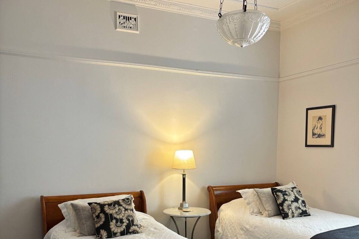 A hotel room with two single beds, a small table, a lamp, and a framed picture on the wall.