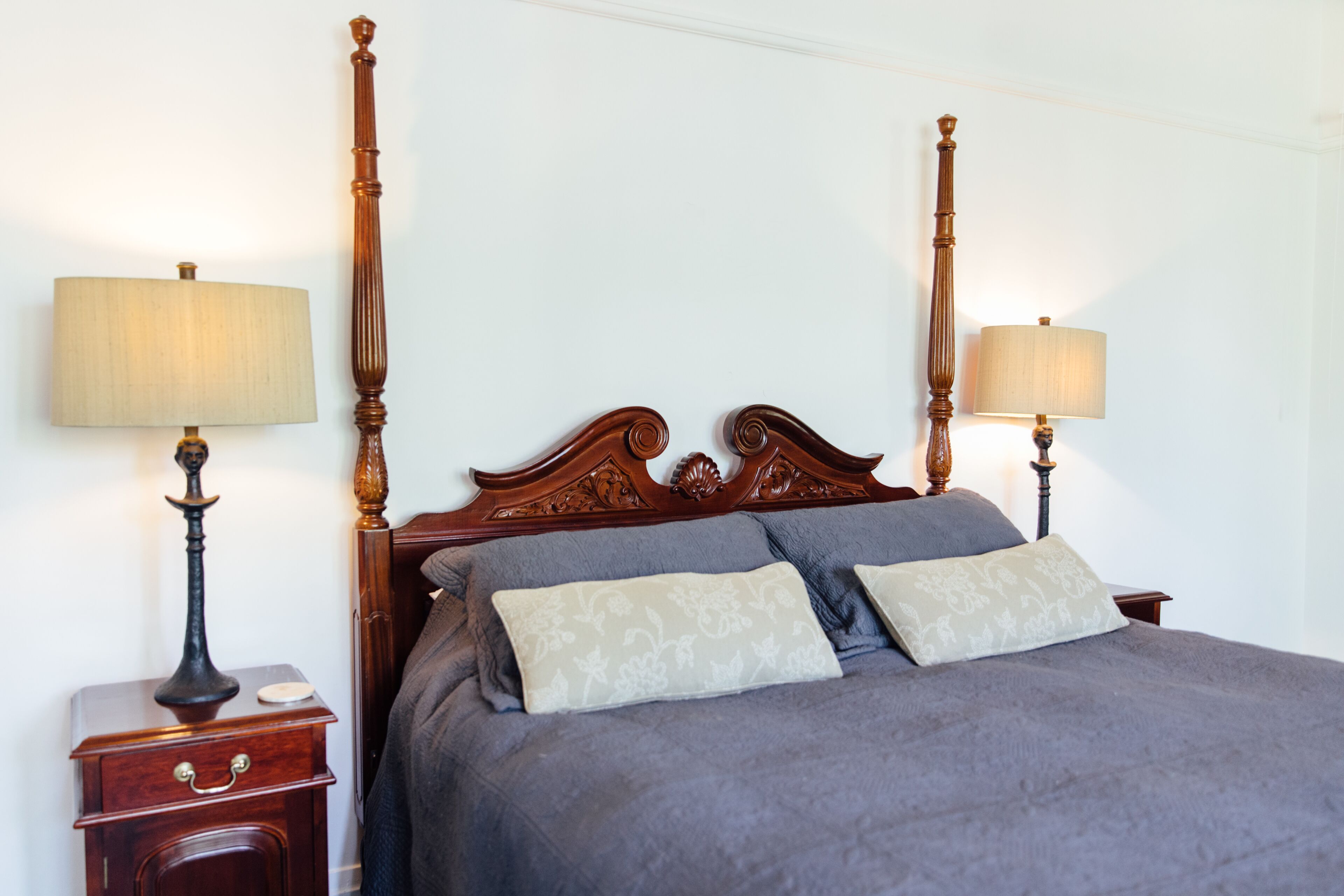 Una cama con dosel, un cabecero de madera, dos mesitas de noche con lámparas y una mesita auxiliar con un cajón.