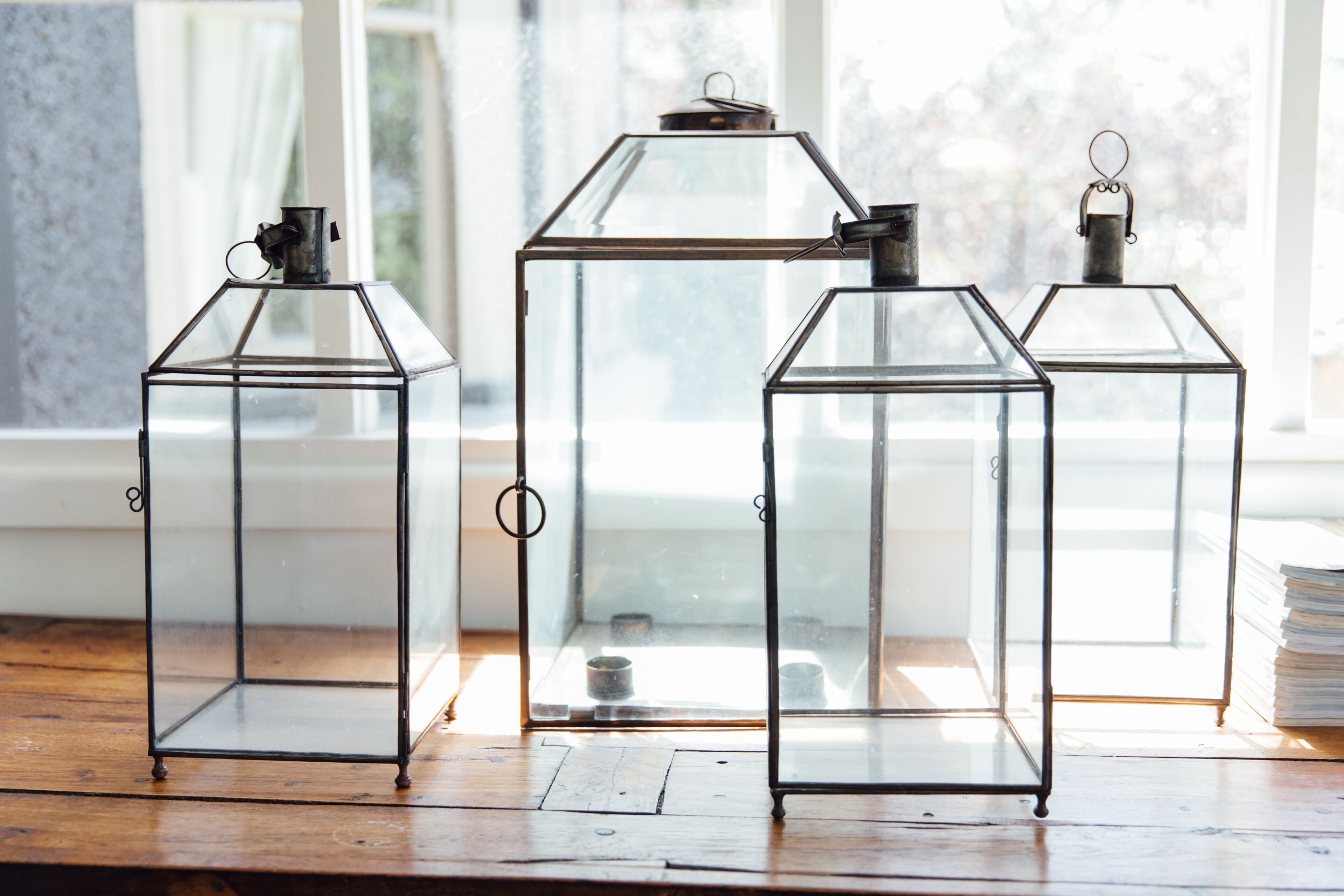 Trois lanternes en verre sont posées sur un plancher en bois, près d’une fenêtre.