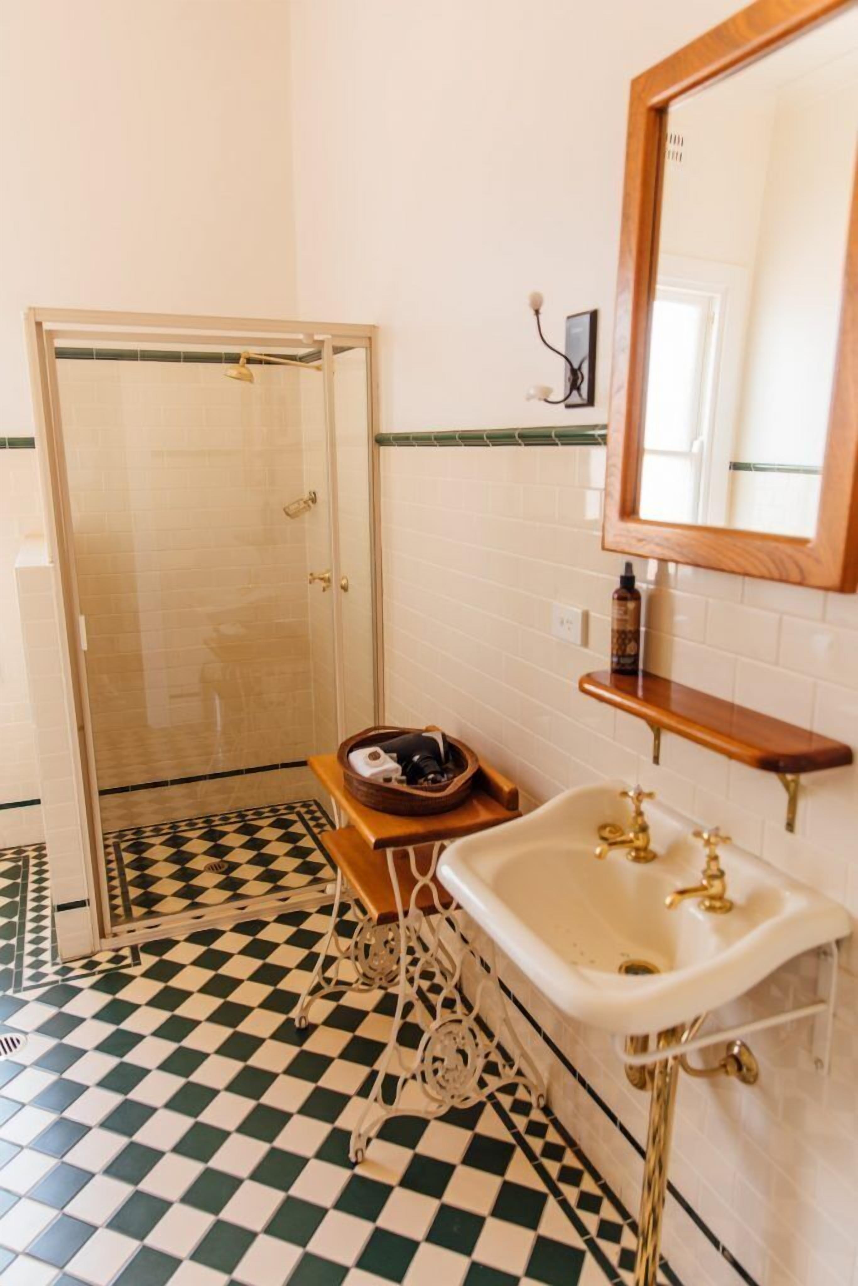A bathroom with a checkered floor, a white sink with gold fixtures, a wooden shelf, and a glass shower enclosure.