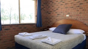 A room with a brick wall, a wooden headboard, a bed with a patterned bedspread, and a window with curtains. - Bullanginya Lodge (Barooga)