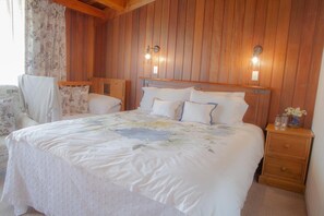 A bedroom with a large bed, two bedside tables, a chair, and wooden paneling on the walls. - Okoroire Hot Springs Hotel (Tapapa)