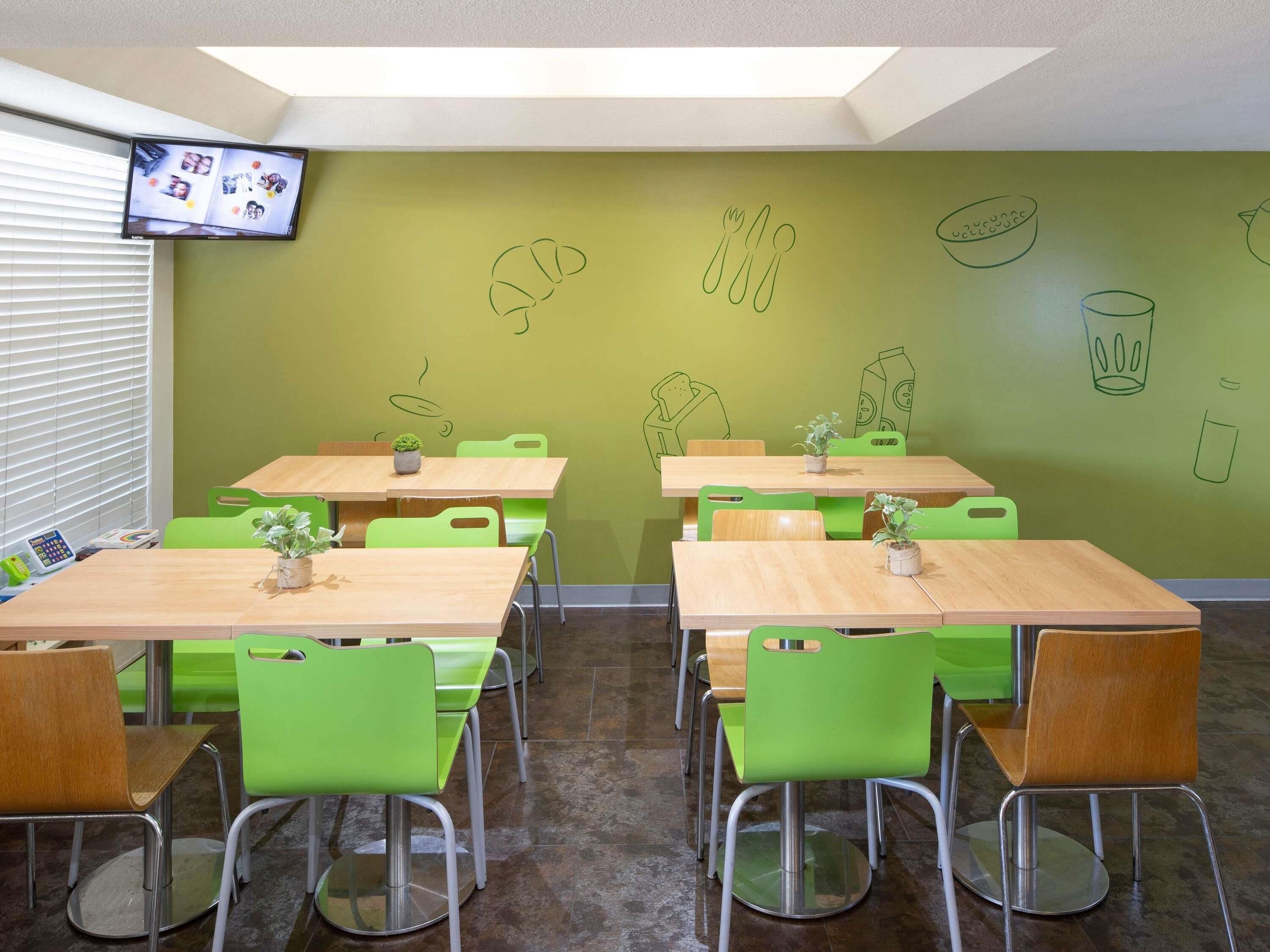 Un espace repas avec des chaises vertes et des tables en bois, un téléviseur fixé au mur et des œuvres d’art murales représentant des aliments.