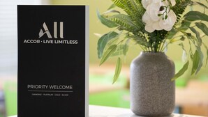 A black brochure titled "ACCOR - LIVE LIMITLESS" with "PRIORITY WELCOME" and "DIAMOND - PLATINUM - GOLD - SILVER" on a table next to a vase with white flowers and green leaves. - Ibis Budget Newcastle (Wallsend)