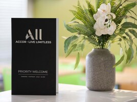 A black brochure titled "ACCOR - LIVE LIMITLESS" with "PRIORITY WELCOME" and "DIAMOND - PLATINUM - GOLD - SILVER" on a table next to a vase with white flowers and green leaves.