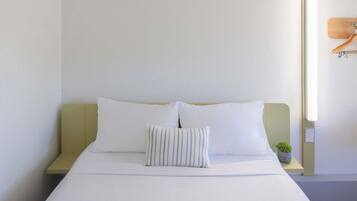A neatly made bed with white linens and two pillows, a small potted plant on a bedside table, and a wall-mounted light fixture.