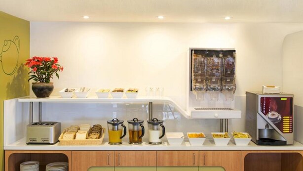 A breakfast bar with various food items, a coffee machine, and a beverage dispenser. - Ibis Budget Newcastle (Wallsend)