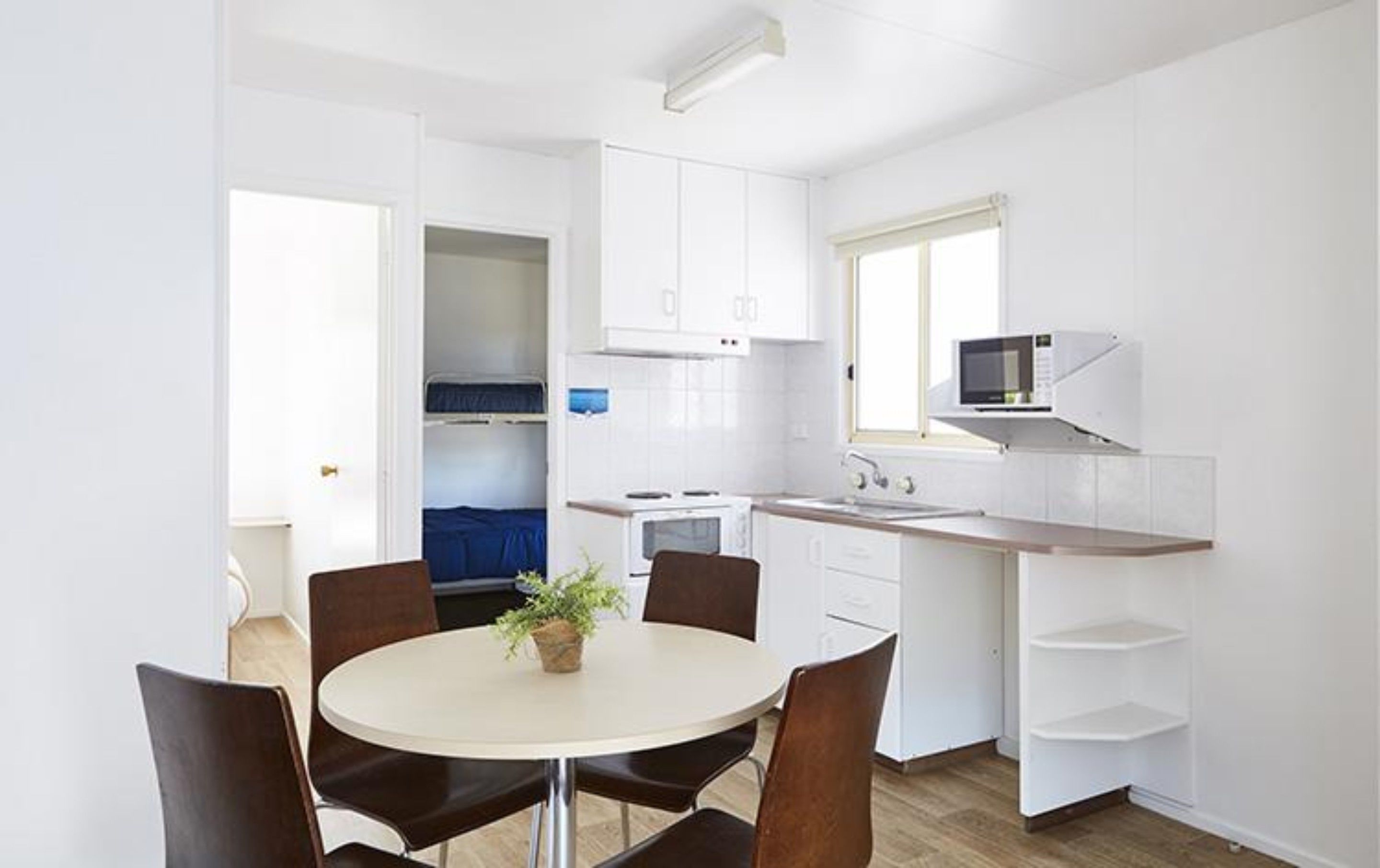 A compact kitchen with white cabinets, a microwave, and a sink. There is a dining area with a round table and chairs, and a small plant on the table.
