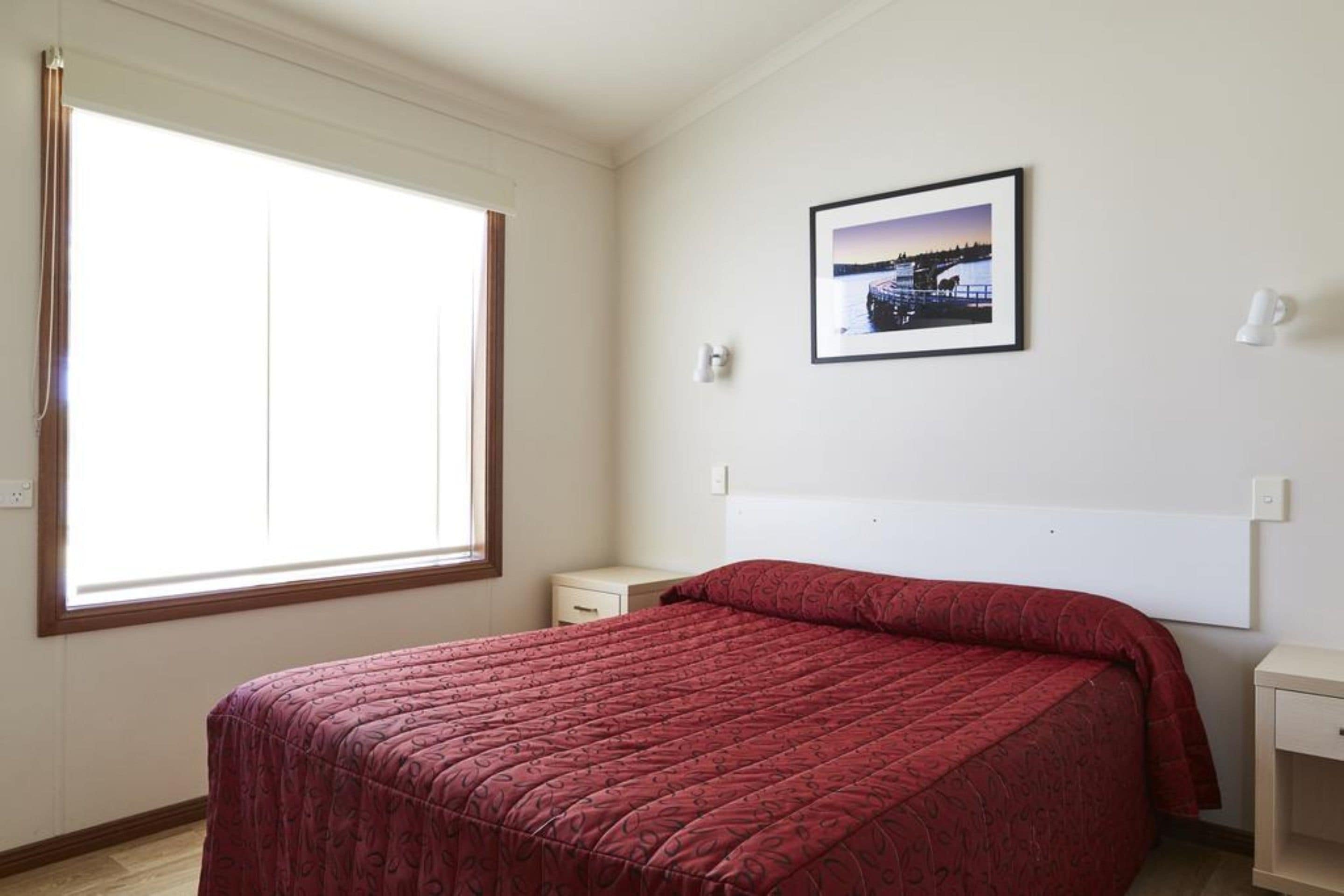 Een slaapkamer met een groot raam, een bed met een rode sprei, een nachtkastje en een klein bijzettafeltje, plus een lijst met een foto aan de muur.