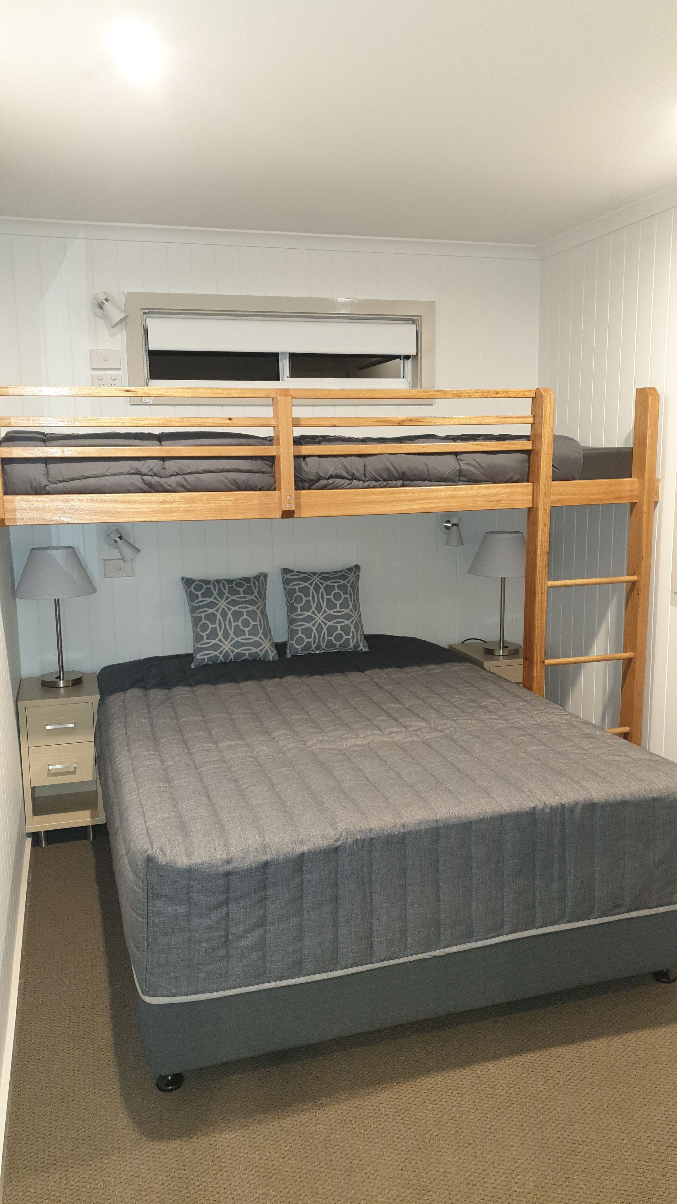 A bunk bed with a grey mattress, two patterned pillows, a bedside table with a lamp, and a window with blinds.