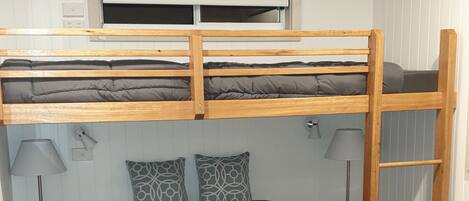 A bunk bed with a grey mattress, two patterned pillows, a bedside table with a lamp, and a window with blinds.