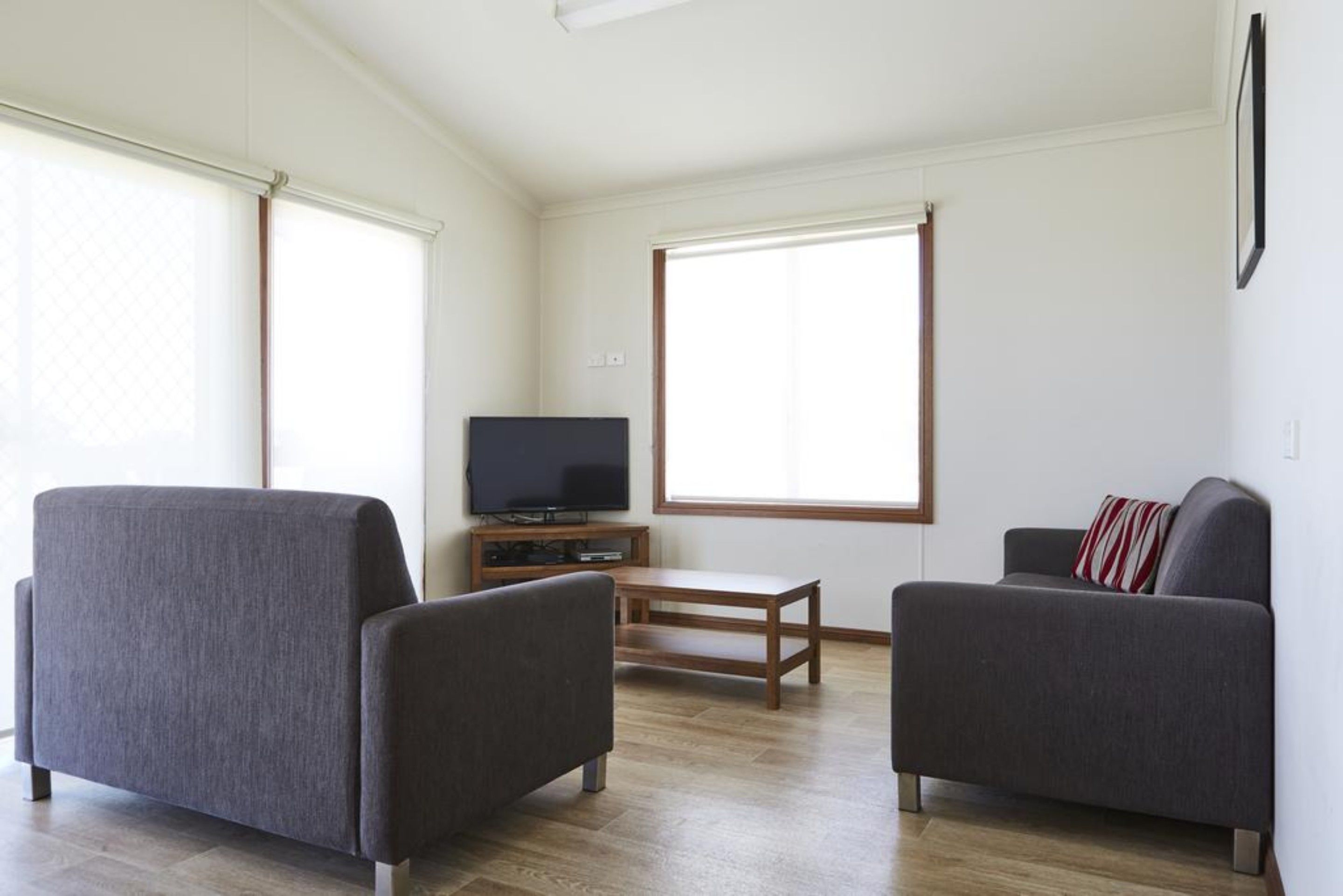 A living room with two grey sofas, a wooden coffee table, a flat-screen TV on a wooden stand, and large windows with blinds.