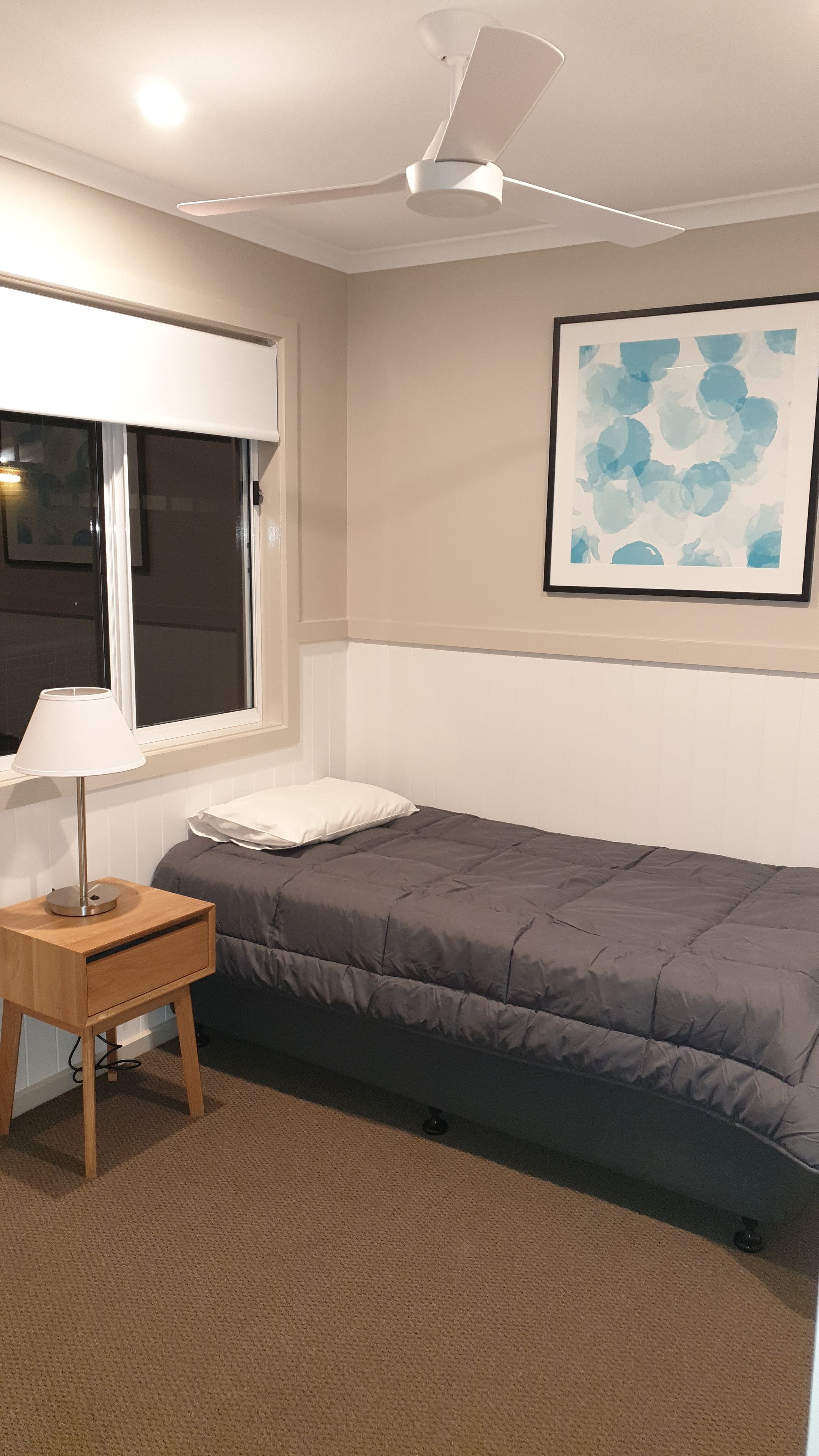Une chambre à coucher comprenant un lit, une table de chevet avec une lampe, un ventilateur de plafond, une fenêtre et un tableau abstrait accroché au mur.