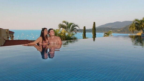 Una pareja en una piscina infinita transparente con vistas al mar y a las montañas.