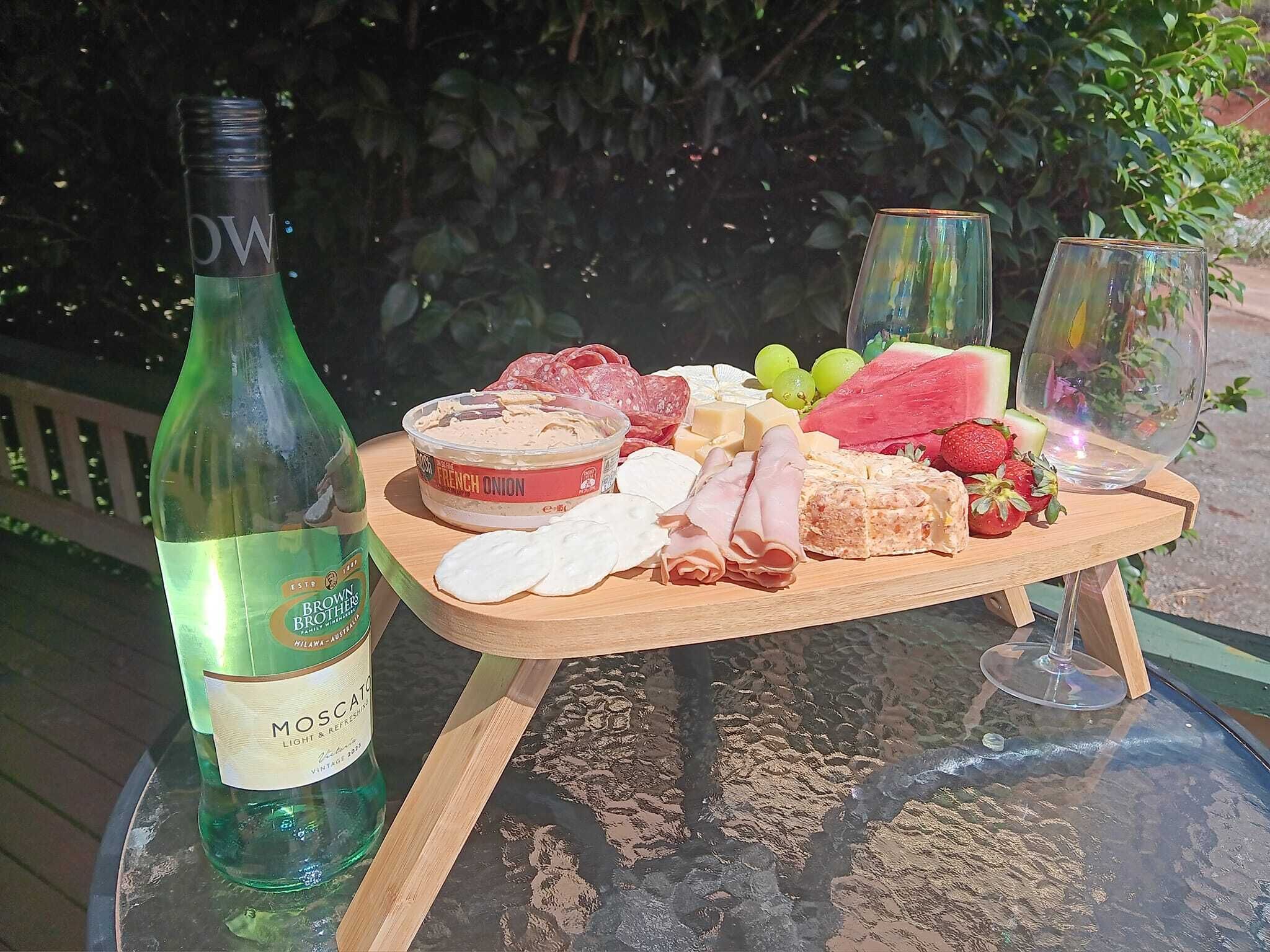 Una mesa de vidrio con una botella de vino, una tabla de madera con varios quesos, embutidos y frutas, y dos copas de vino.