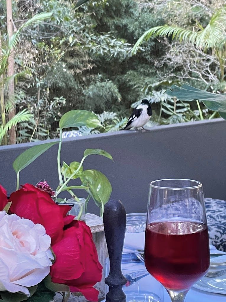 A table with a glass of red liquid, a vase with red and white roses, and a bird perched on a railing.
