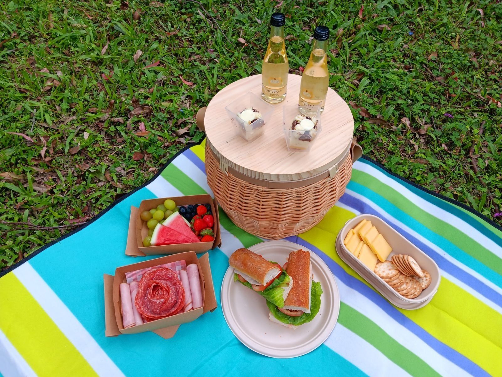 Ein Picknick mit einem Korbwesen, Flaschen, einem Sandwich, Obst, Käse und Salami auf einer Streifen-Decke.
