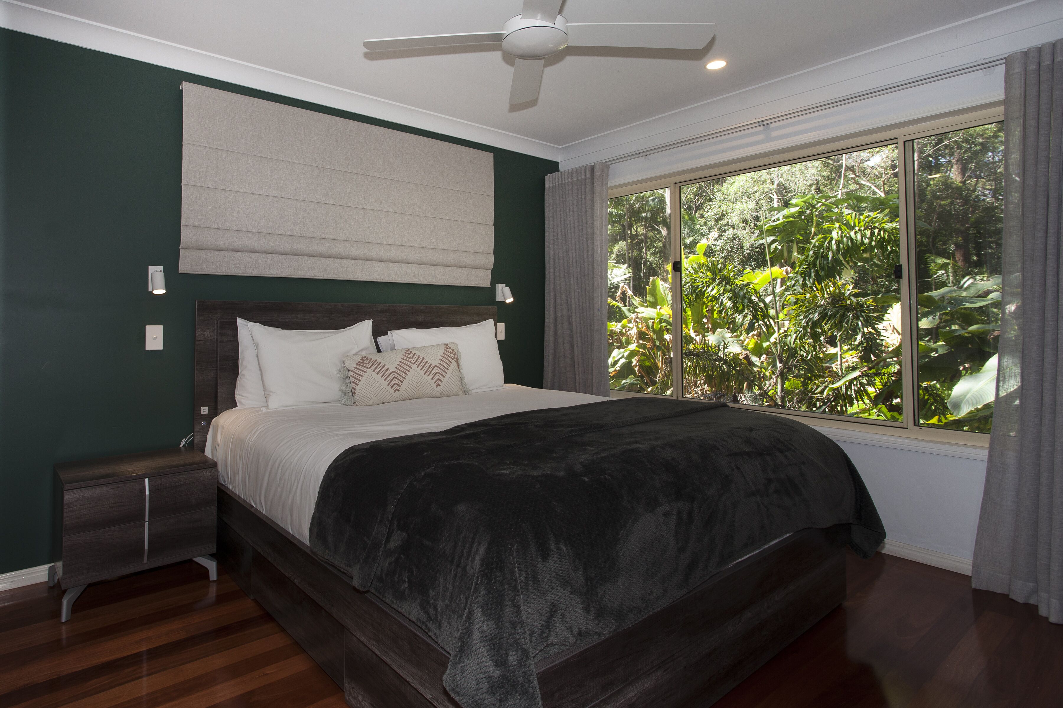A bedroom with a large bed, a nightstand, a ceiling fan, and a window with a view of greenery.