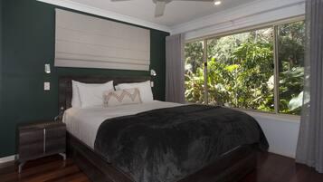 A bedroom with a large bed, a nightstand, a ceiling fan, and a window with a view of greenery.