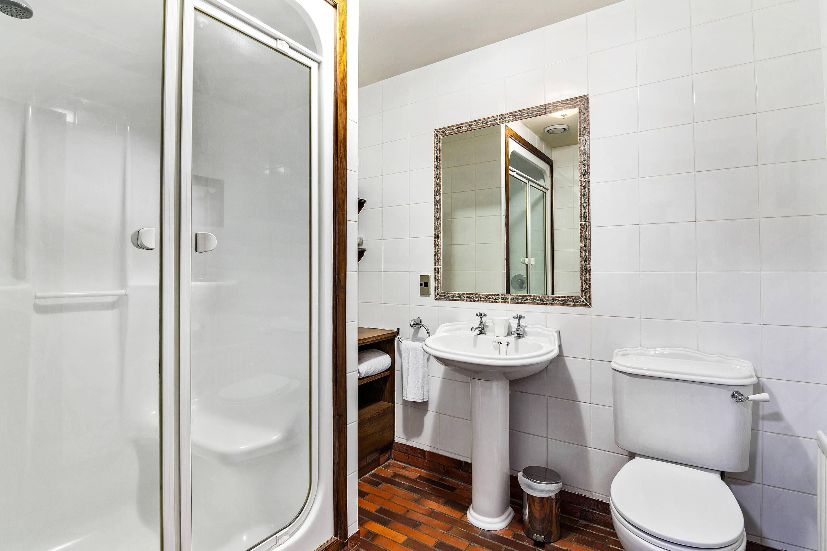 Un baño con cabina de ducha de vidrio, lavamanos pedestal, inodoro y espejo con marco decorativo.