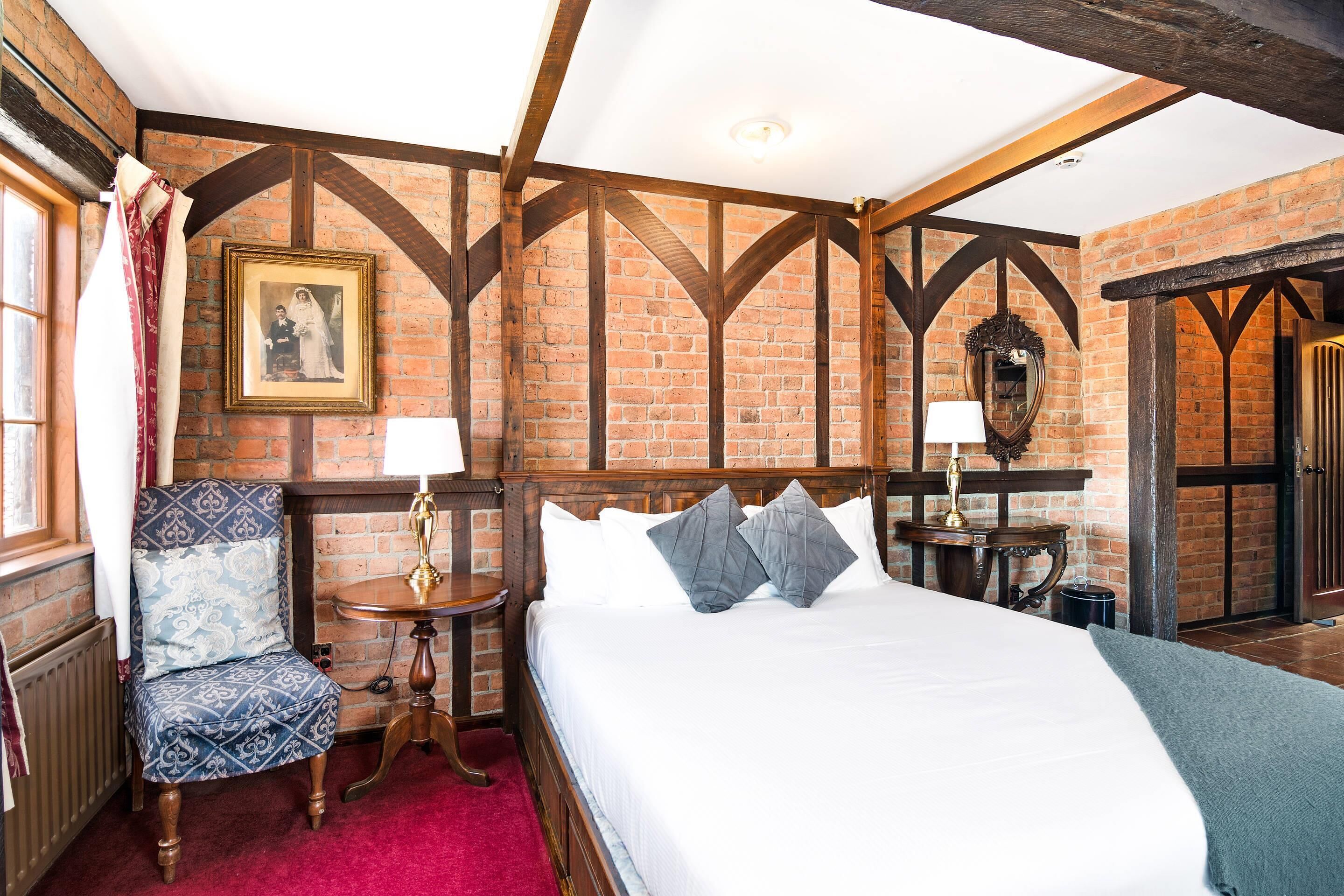 A traditional room with exposed brick walls, a wooden ceiling, a large bed with white linens, a patterned armchair, and a small table with a lamp.