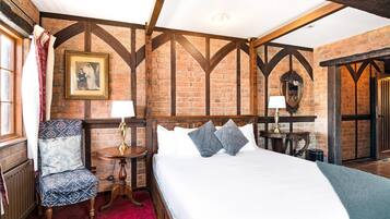A traditional room with exposed brick walls, a wooden ceiling, a large bed with white linens, a patterned armchair, and a small table with a lamp.