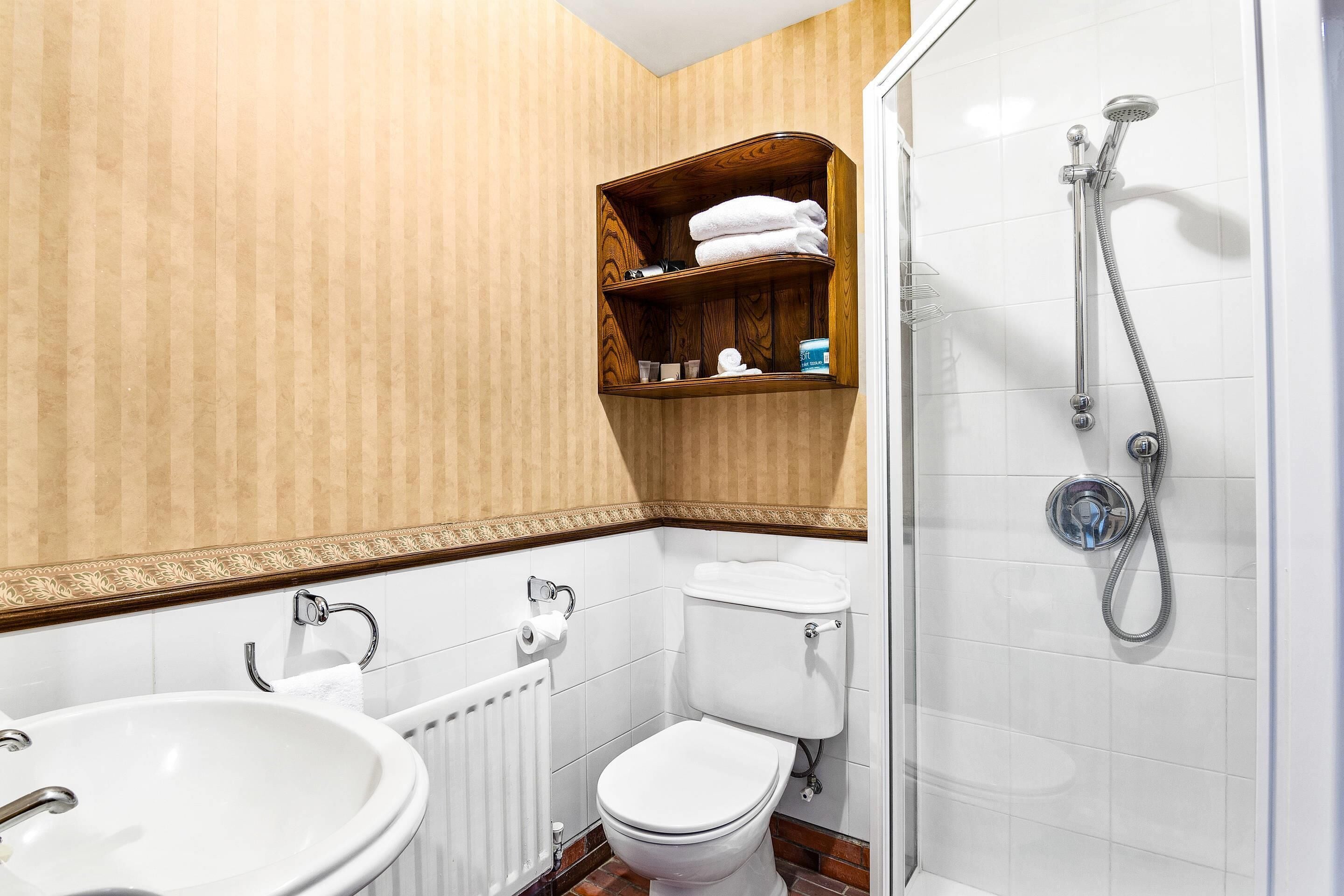 Un baño con lavamanos, inodoro y ducha blancos. Hay un estante de madera sobre el inodoro con toallas y artículos de aseo.