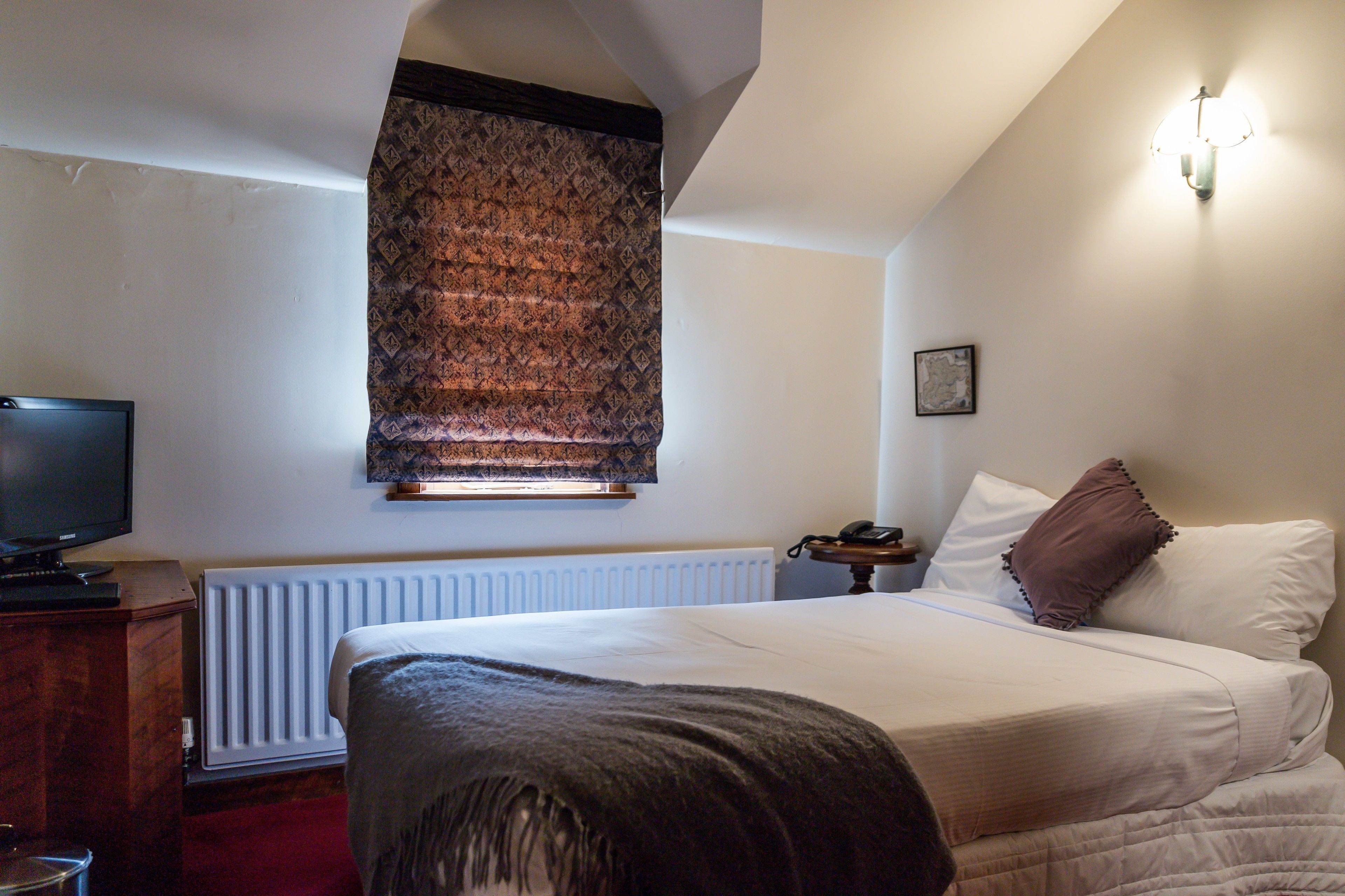A bedroom with a bed, a television, a radiator, and a window with a patterned blind.