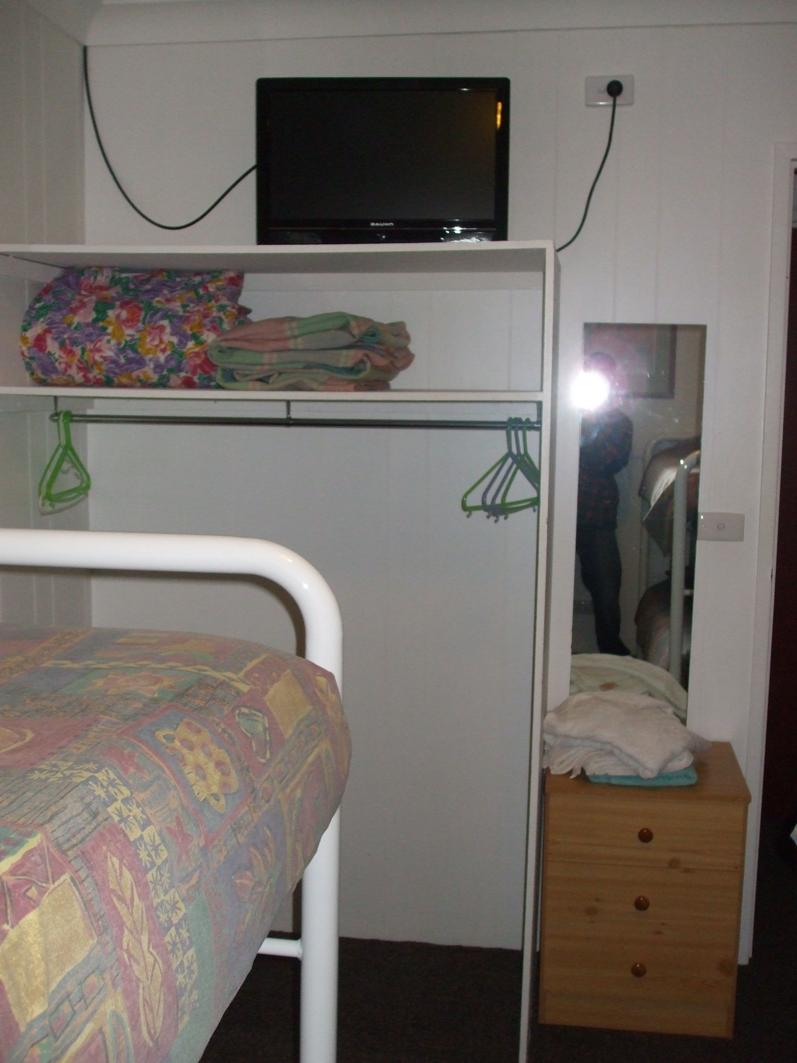 A bedroom with a bed, a television on a shelf, a mirror, and a small wooden dresser.