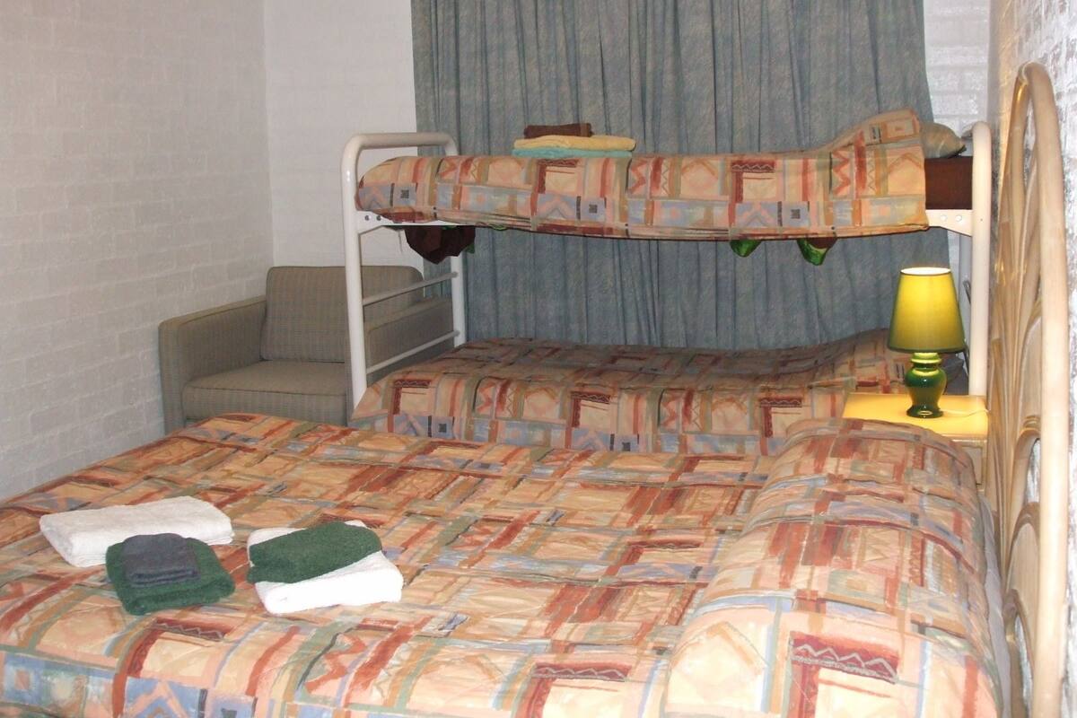 A bunk bed room with a patterned bedspread, a small bedside table with a lamp, and a chair.