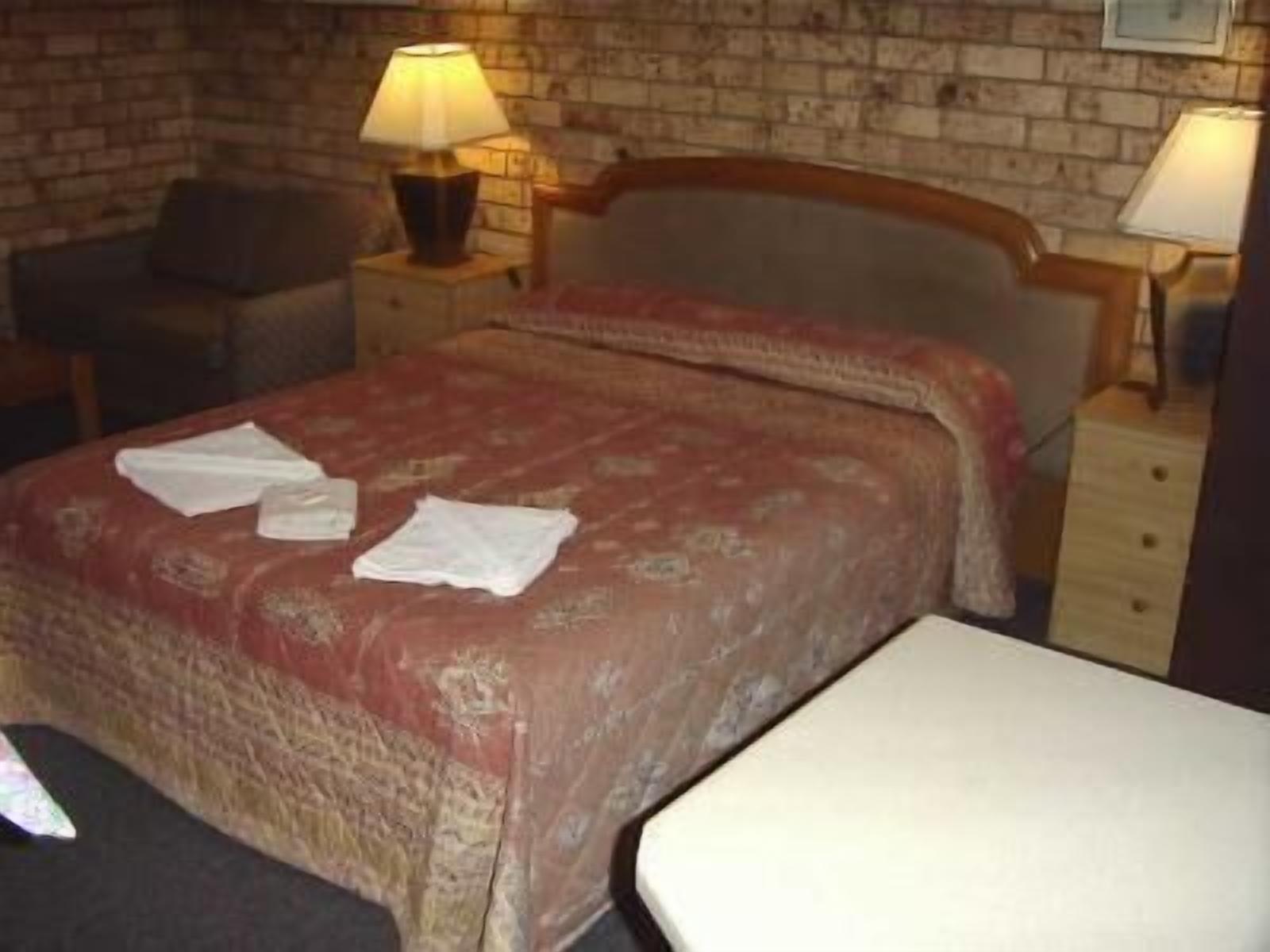 A neatly made bed with a red and beige patterned bedspread, two pillows, and a white bedside table with a lamp.