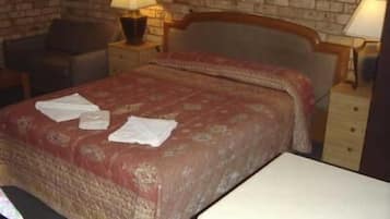 A neatly made bed with a red and beige patterned bedspread, two pillows, and a white bedside table with a lamp.
