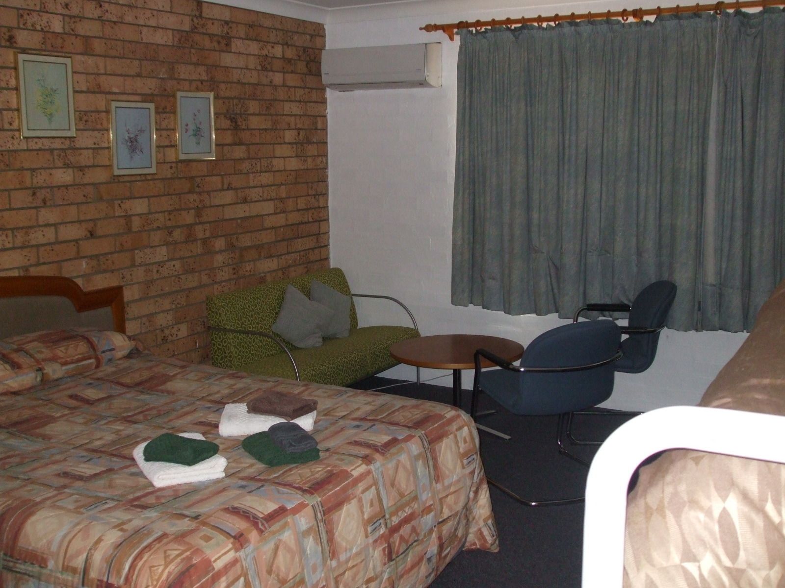 A hotel room with a brick wall, a bed with a patterned bedspread, a green sofa, a small round table, a blue chair, and a window with curtains.