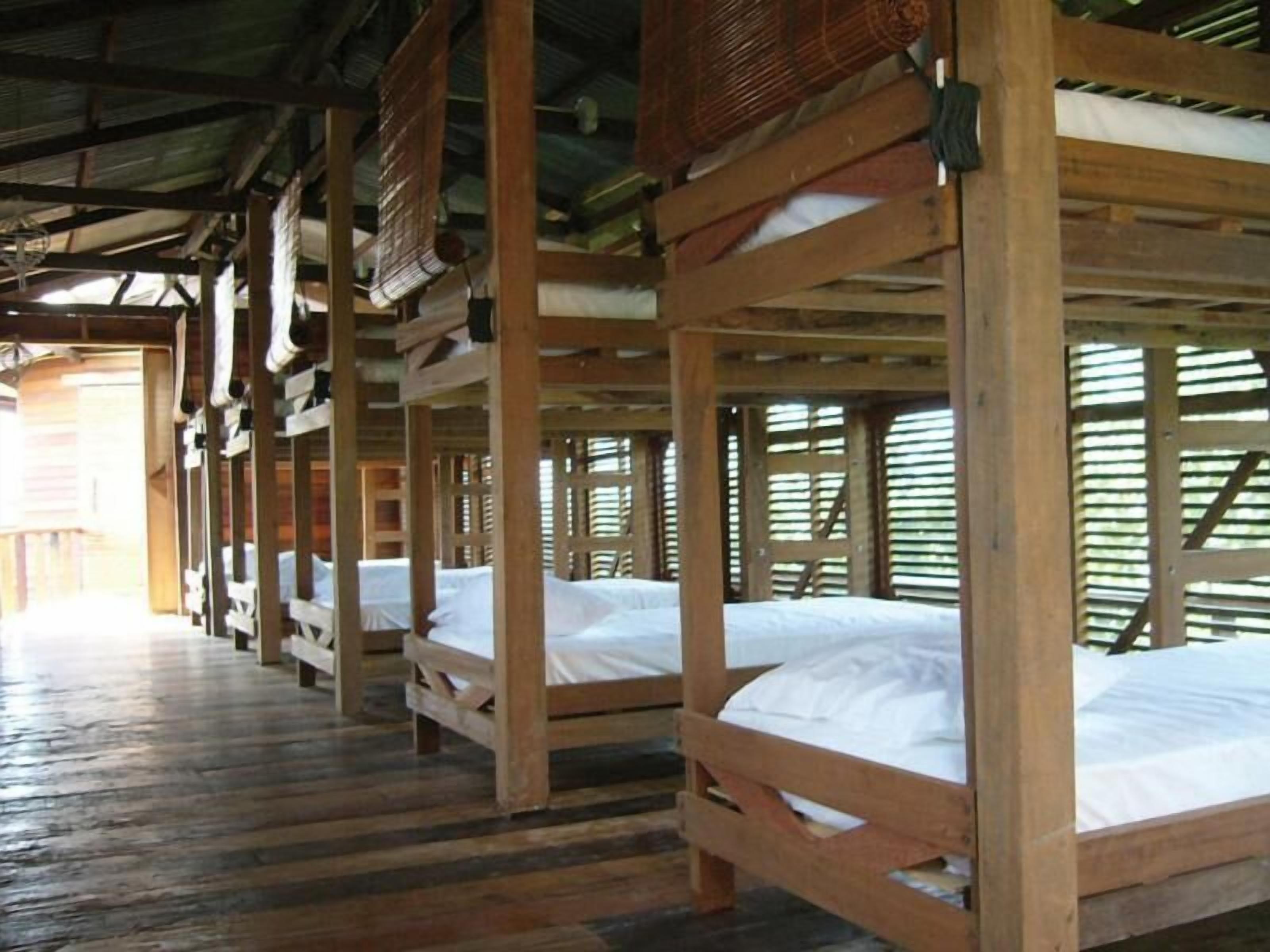 Varias literas de madera con ropa de cama blanca, bajo una estructura cubierta con piso de madera.