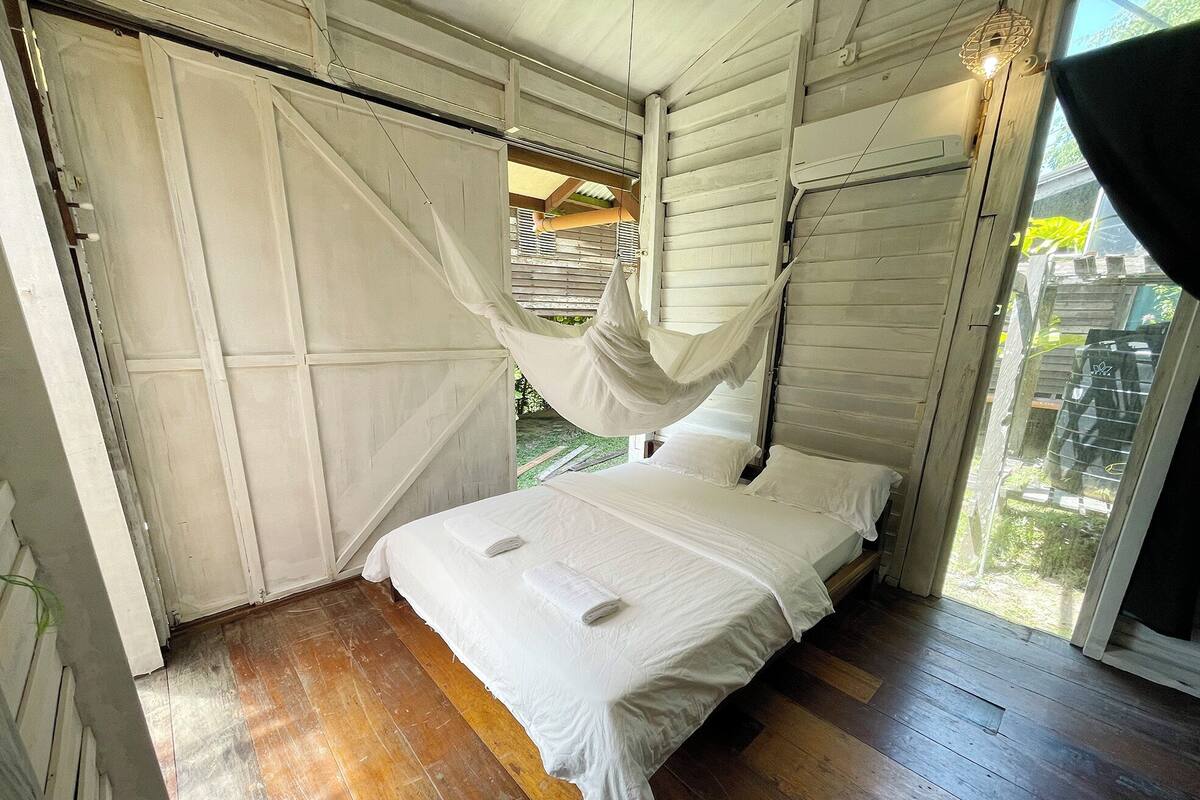 A bedroom with a bed, wooden flooring, a canopy, and a view of the outdoors.
