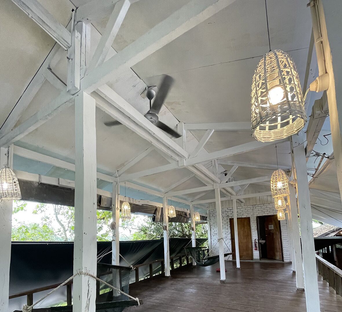 A covered outdoor area with white beams and a ceiling fan, featuring hanging wicker lamps and hammocks.